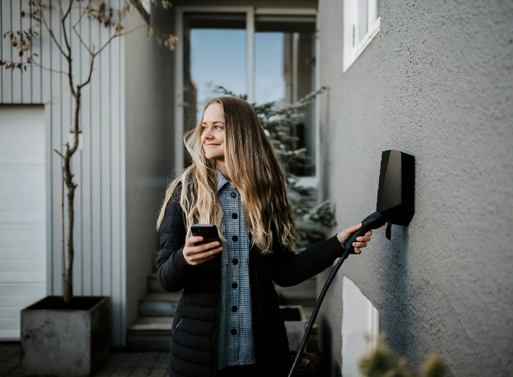 Woman holding smartphone while plugging in black Easee EV charger mounted on exterior wall of modern home